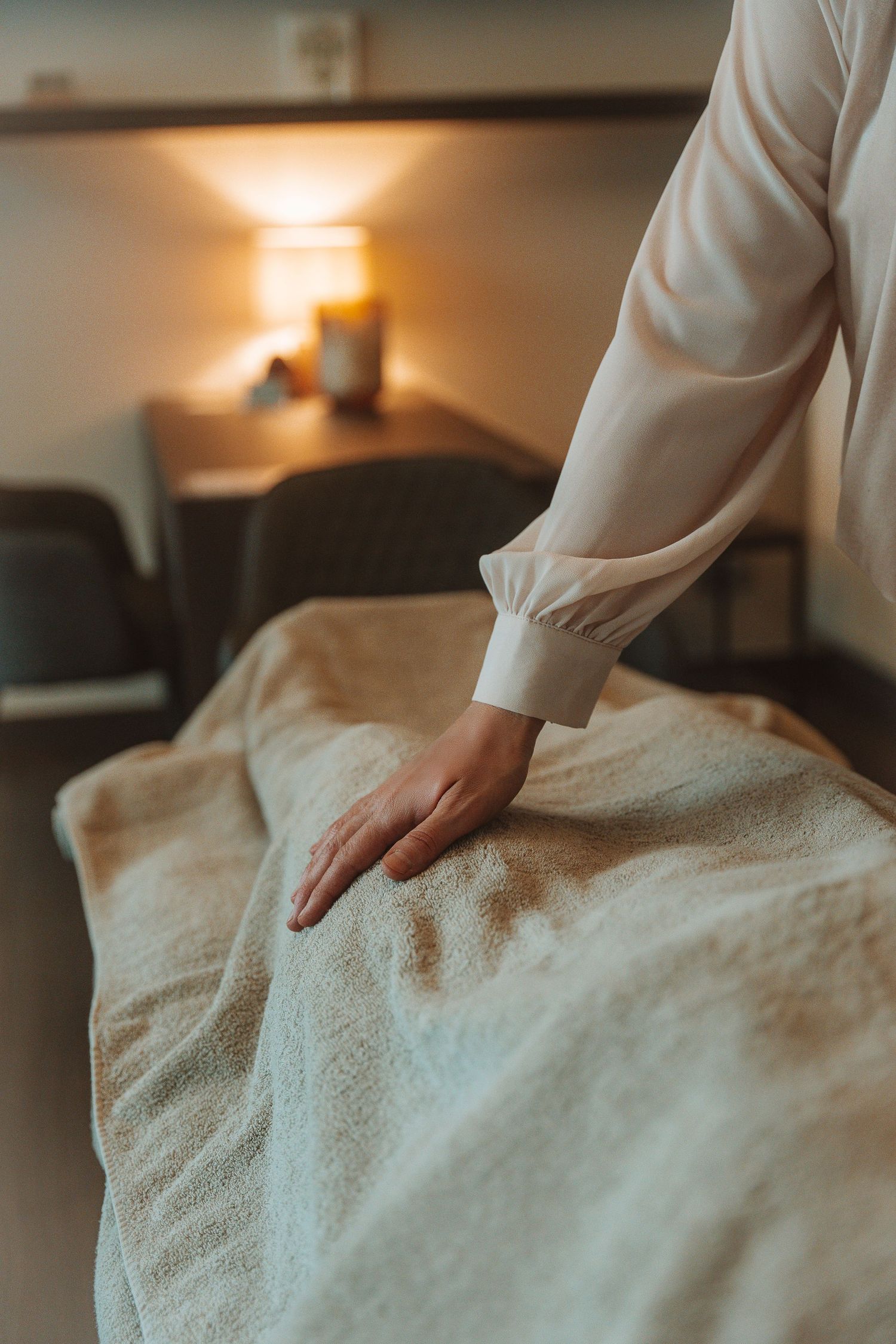 A hand in a white sleeve smooths a beige textured blanket or massage table cover in a dimly lit spa setting.
