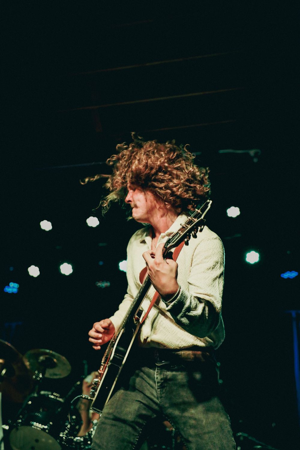 Musician with wild curly hair performing passionately on electric guitar in dark venue.