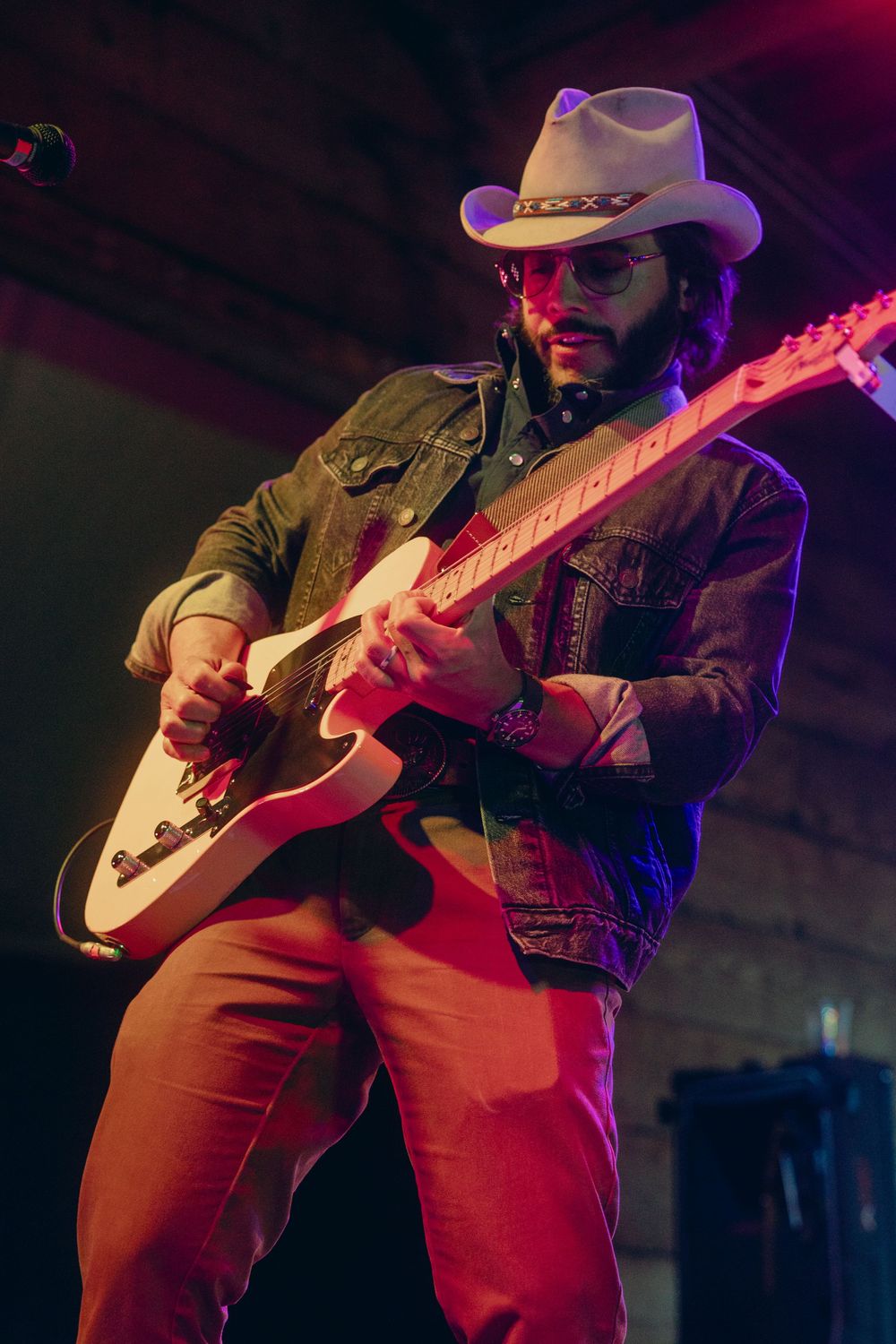 A musician in a cowboy hat plays acoustic guitar under dramatic red and blue stage lighting at a live performance.