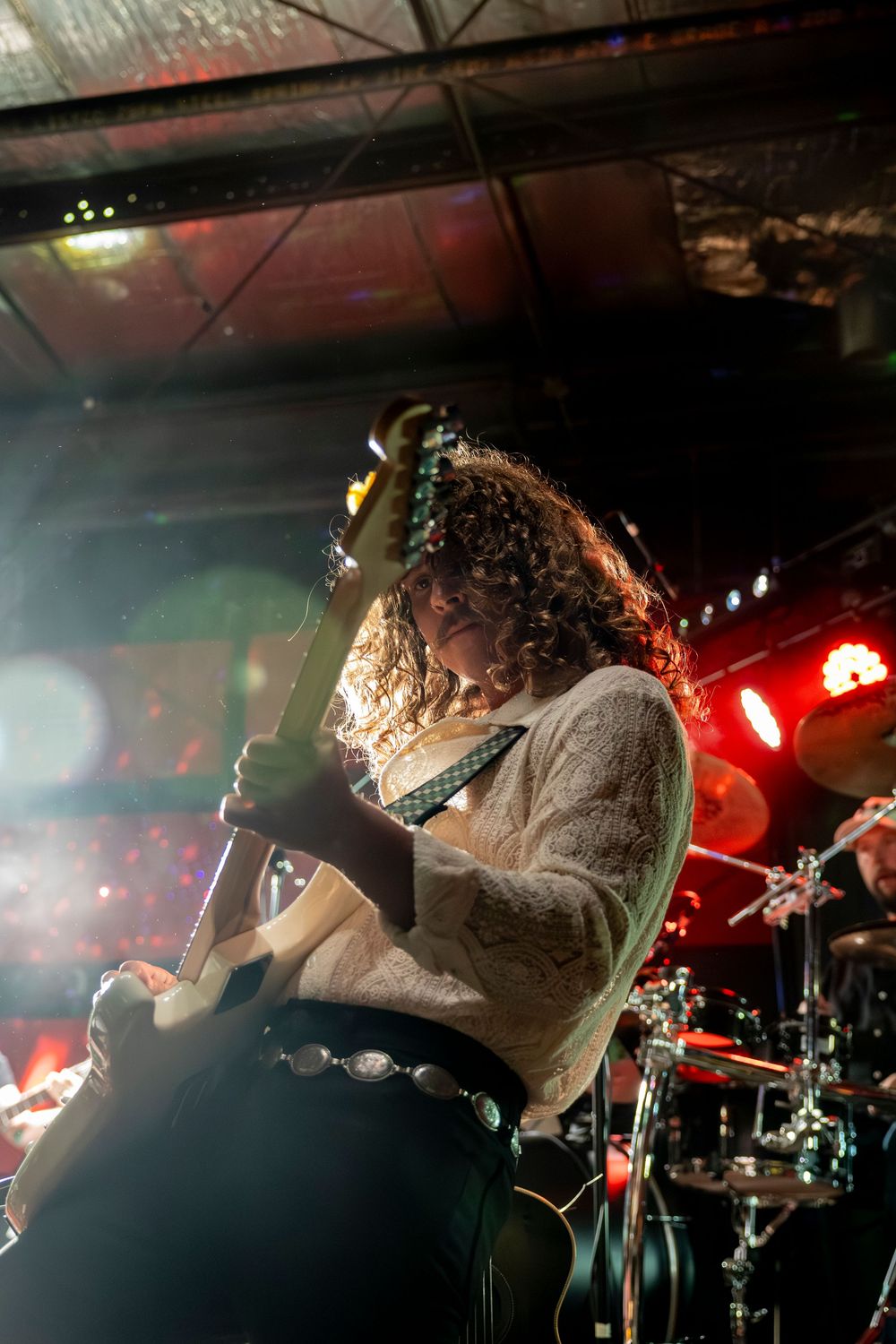 Curly-haired guitarist performs on smoky stage under red lights with drums visible in background.