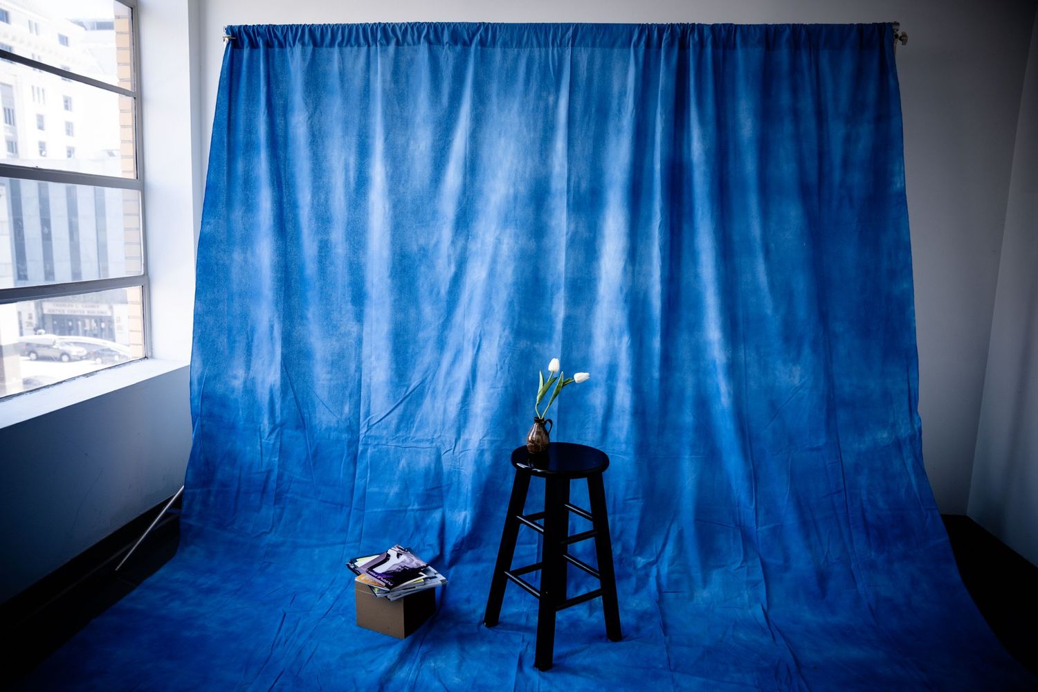 Simple photography studio setup with blue backdrop and wooden stool near window.