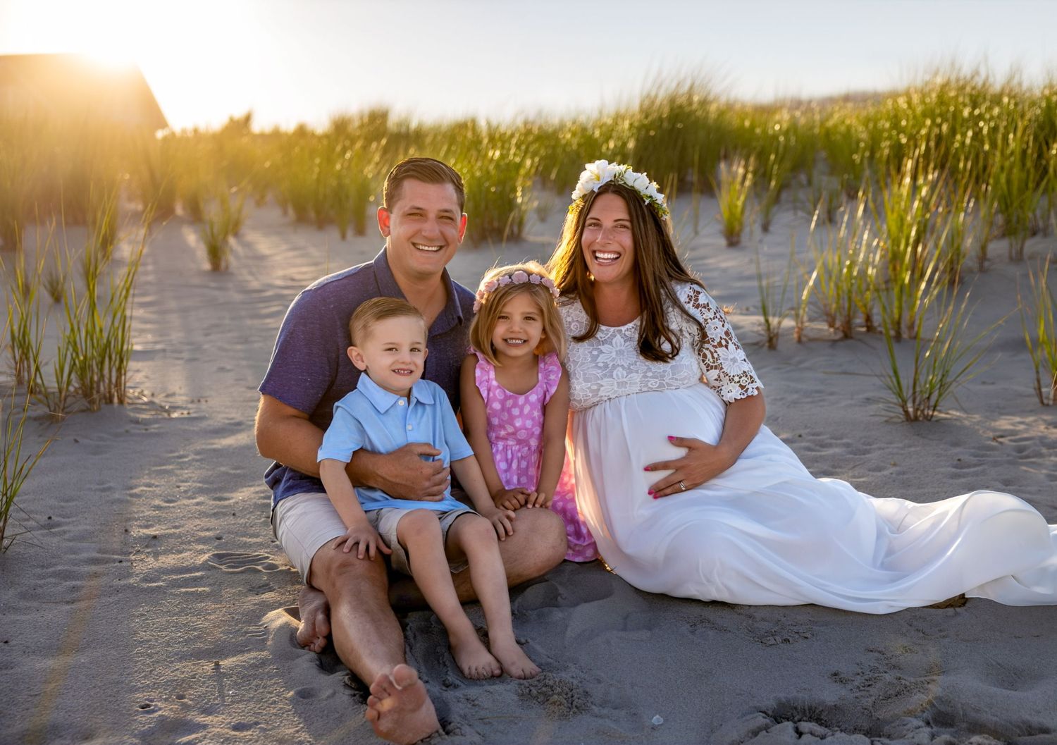 Family sitting together on beach sand at sunset with coastal grass in background.