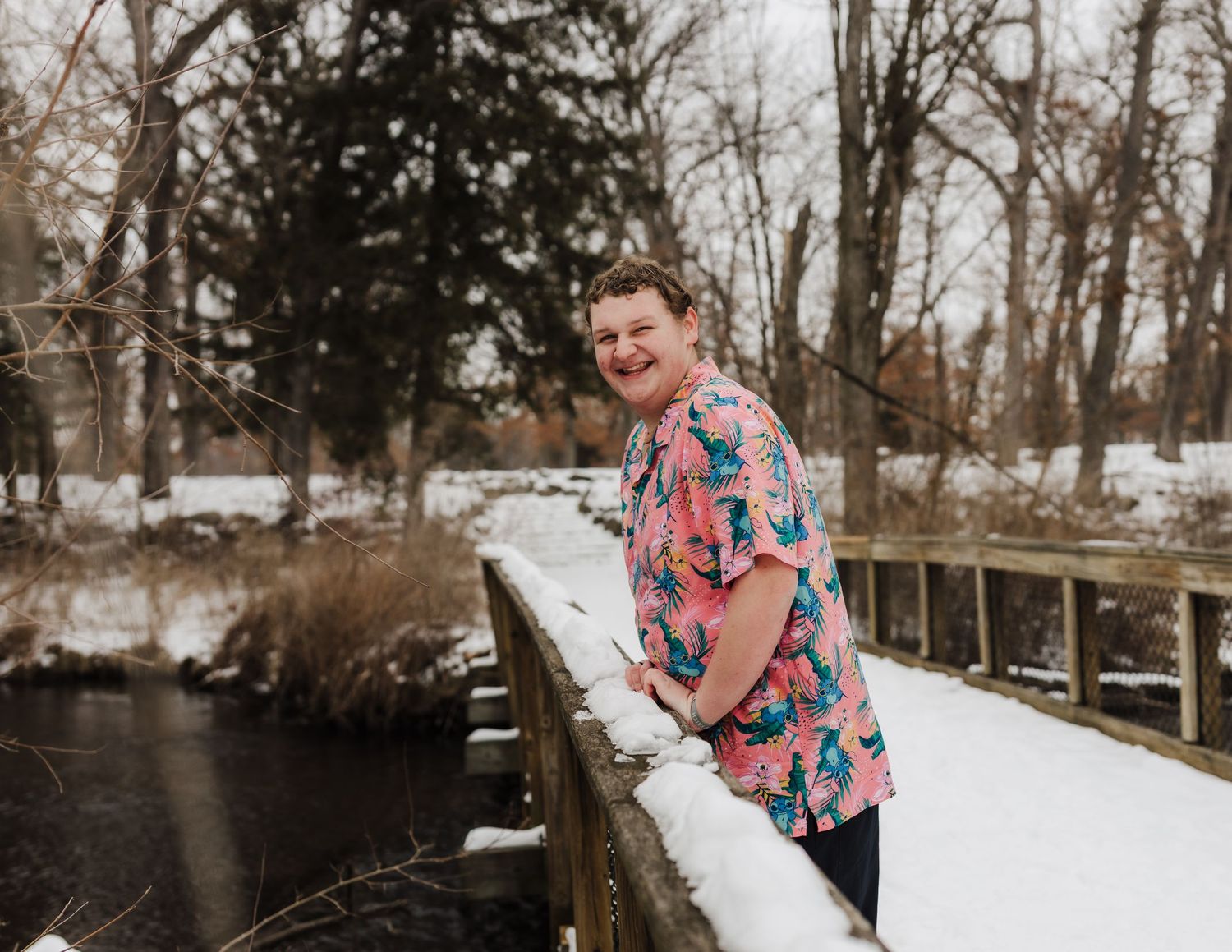 Person in a colorful floral shirt stands on a snowy bridge over a frozen pond during winter.