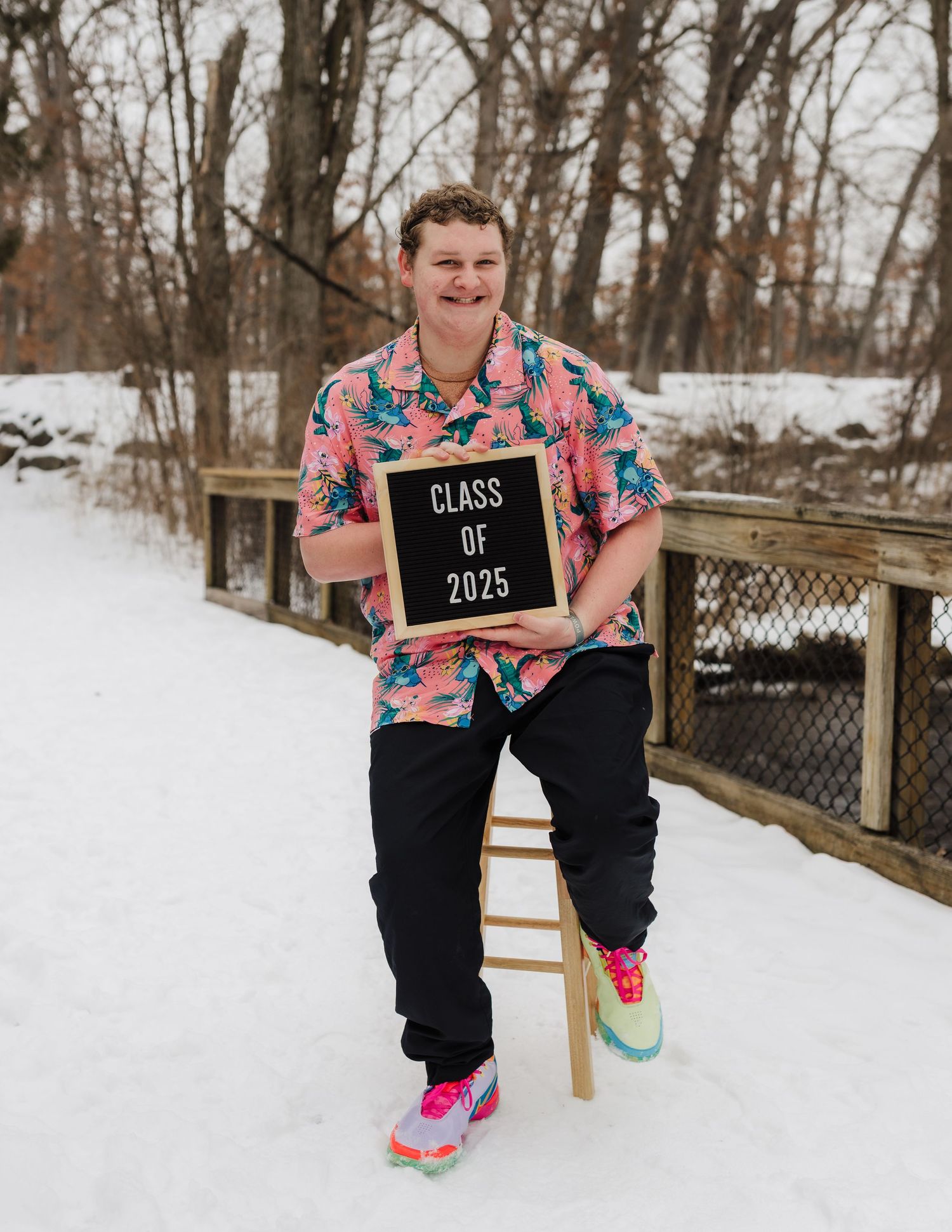 A boy in a tropical print shirt holds a 'Class of 2025' sign while standing in a snowy winter setting with trees.