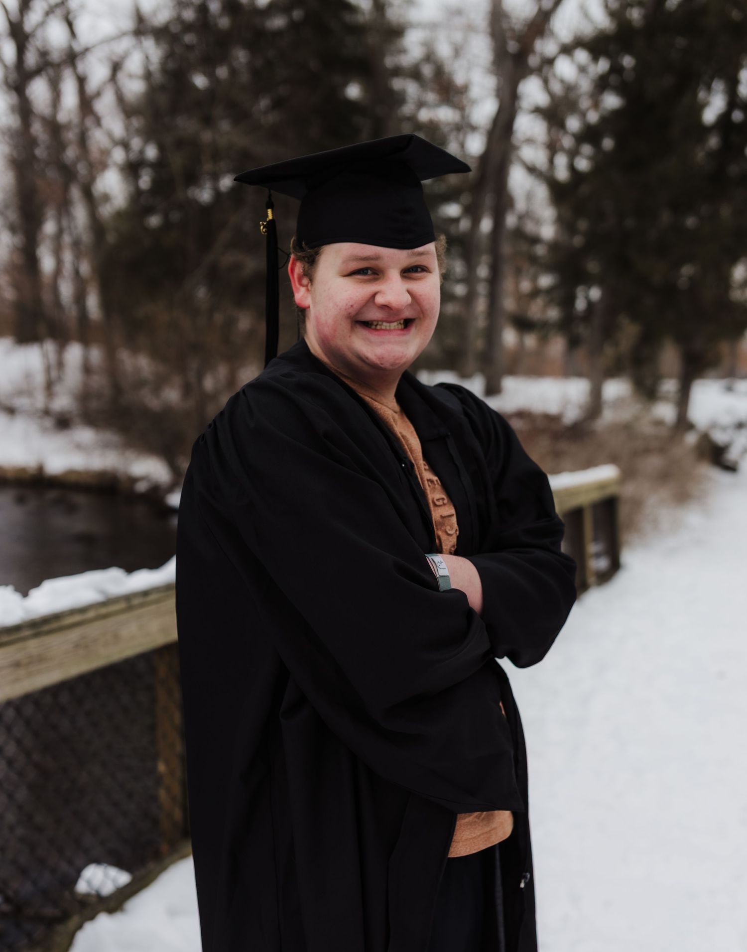 A graduate in black cap and gown poses on a snowy wooden walkway against a winter forest backdrop.
