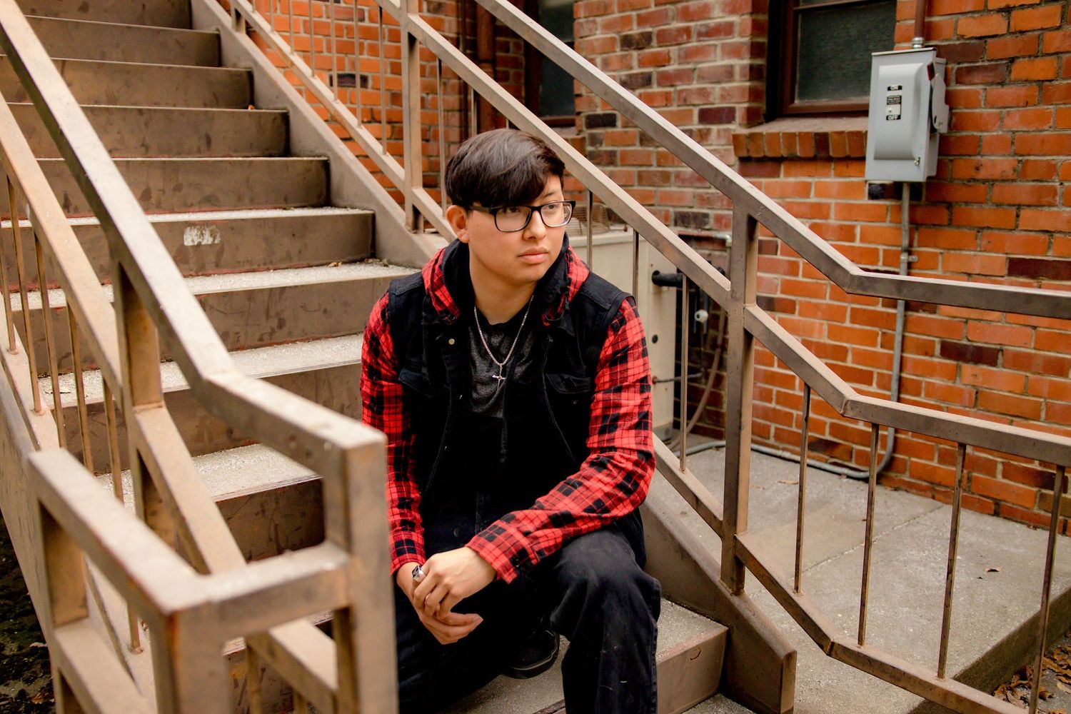 Person in a red plaid flannel and black vest sits on concrete steps outside a brick building.