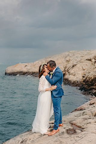 Black and white photo of romantic couple embracing against rocks by the ocean.
