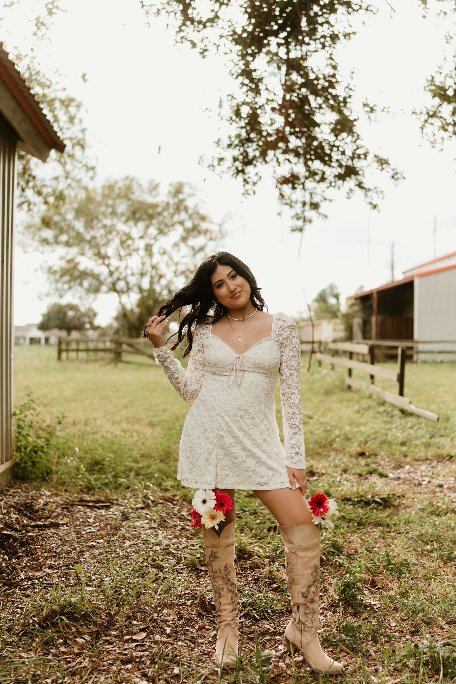 Woman in white lace dress poses in a rustic outdoor setting with trees and fencing.