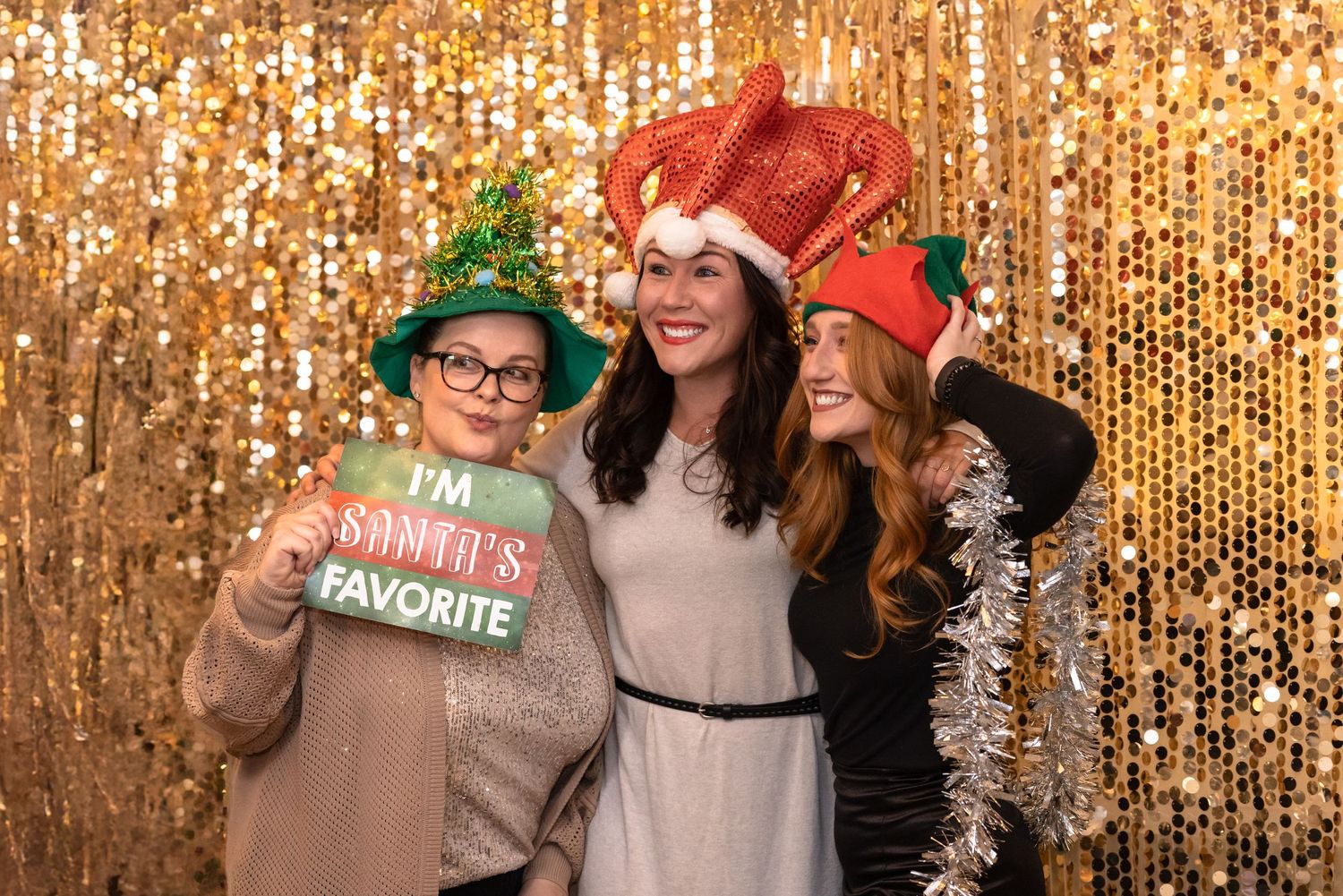 Four friends pose together wearing festive holiday hats against a sparkly gold sequin backdrop at a Christmas party.
