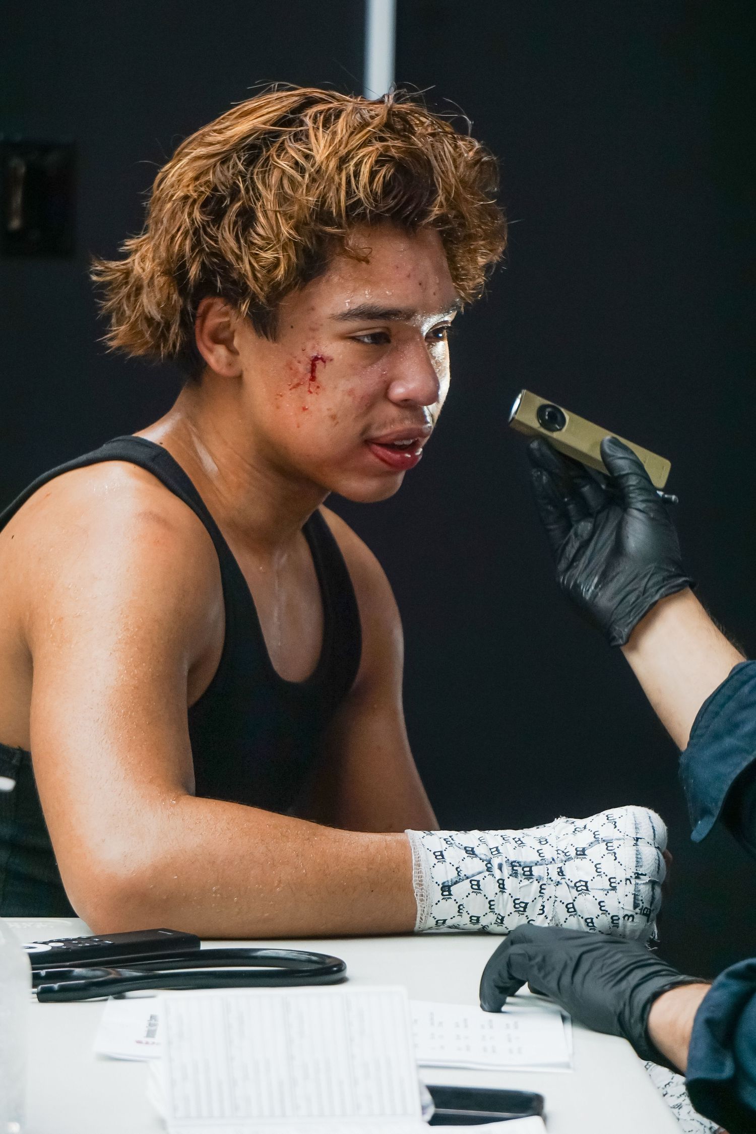 A fighter with blonde hair and facial cuts receives medical attention wearing a black tank top and bandaged hand.