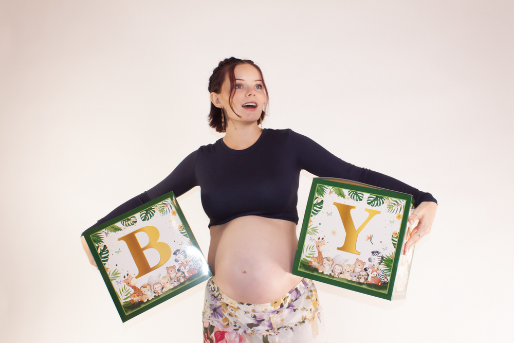 A pregnant woman in a black crop top holds decorative floral baby letter signs while posing against a pink background.