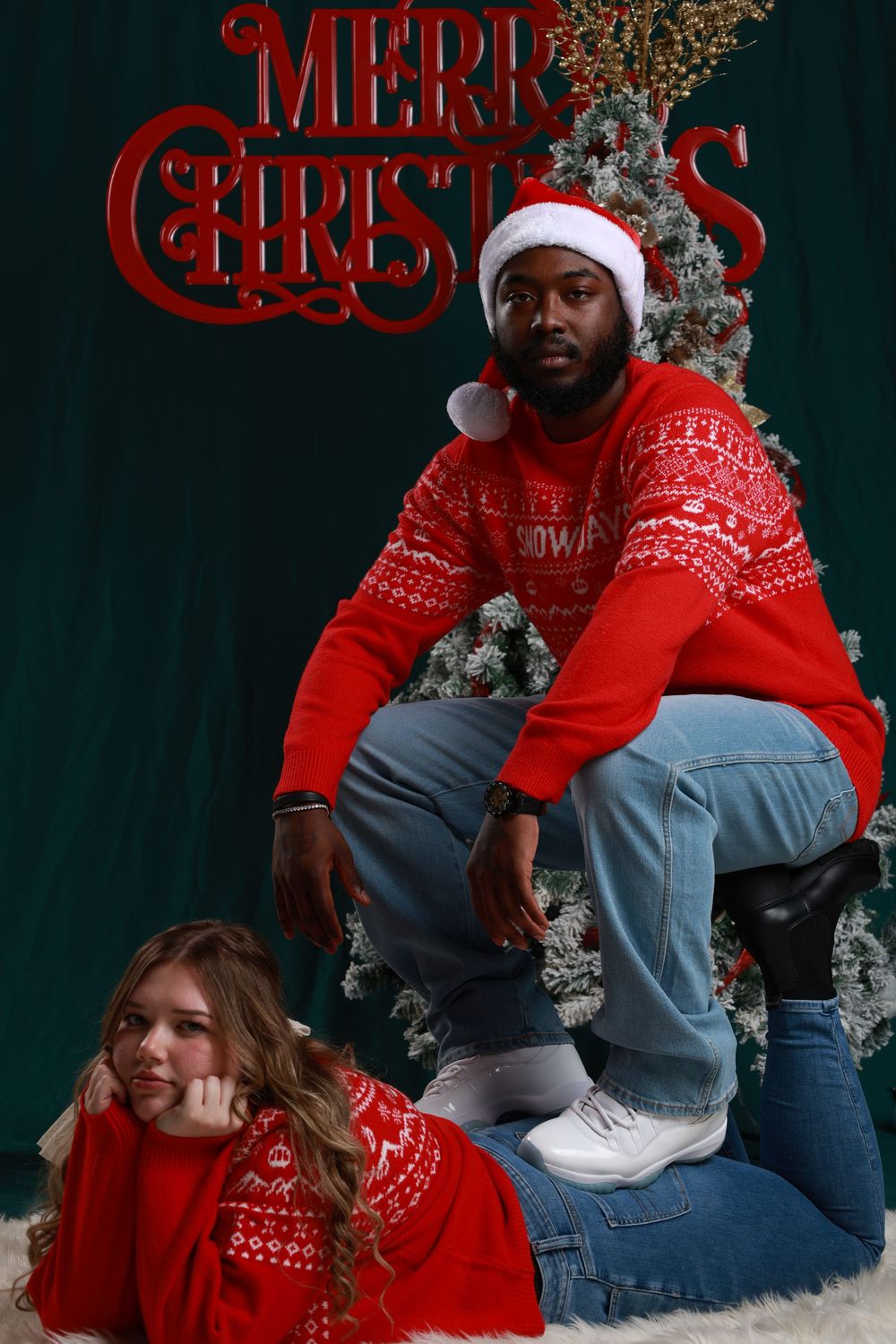 Couple in matching red Christmas sweaters and Santa hats posing in front of a decorated Christmas tree with Merry Christmas sign.