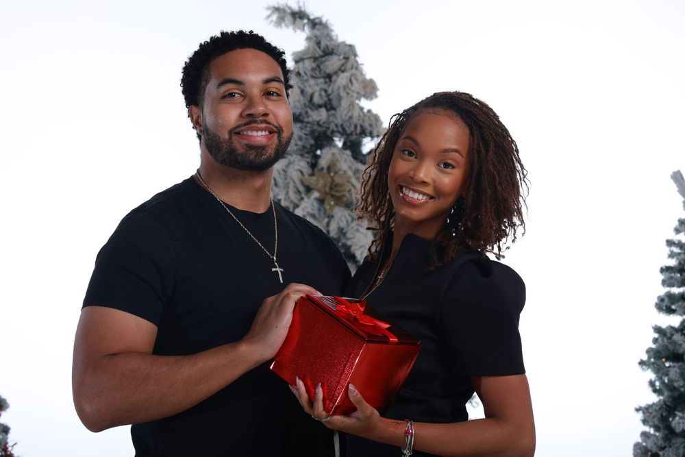 A smiling couple in black outfits holding a red gift box near a snow-dusted Christmas tree.