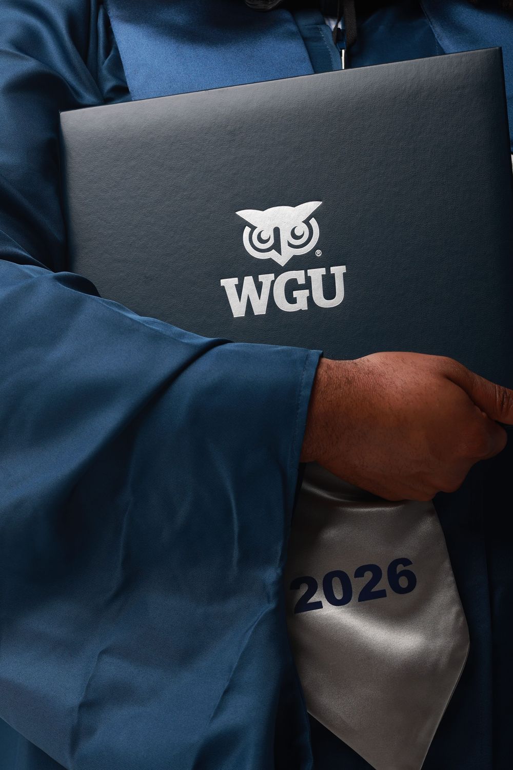 A WGU graduate in blue regalia holds a diploma cover with the university owl logo, with a 2026 sash visible.