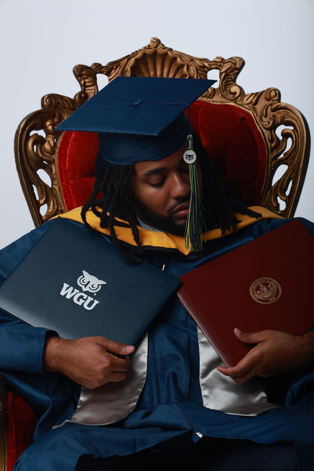 Graduate in blue cap and gown holds two WGU diplomas while resting in ornate red throne chair.