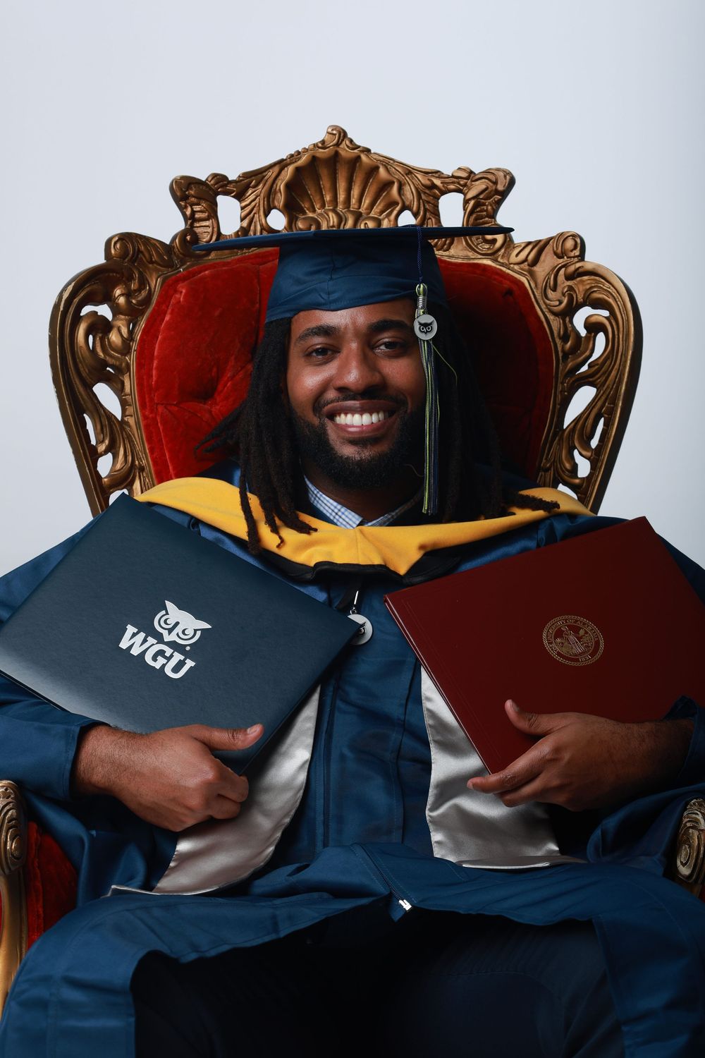 Smiling graduate in blue cap and gown sits on ornate throne chair holding two diplomas, including a WGU degree.