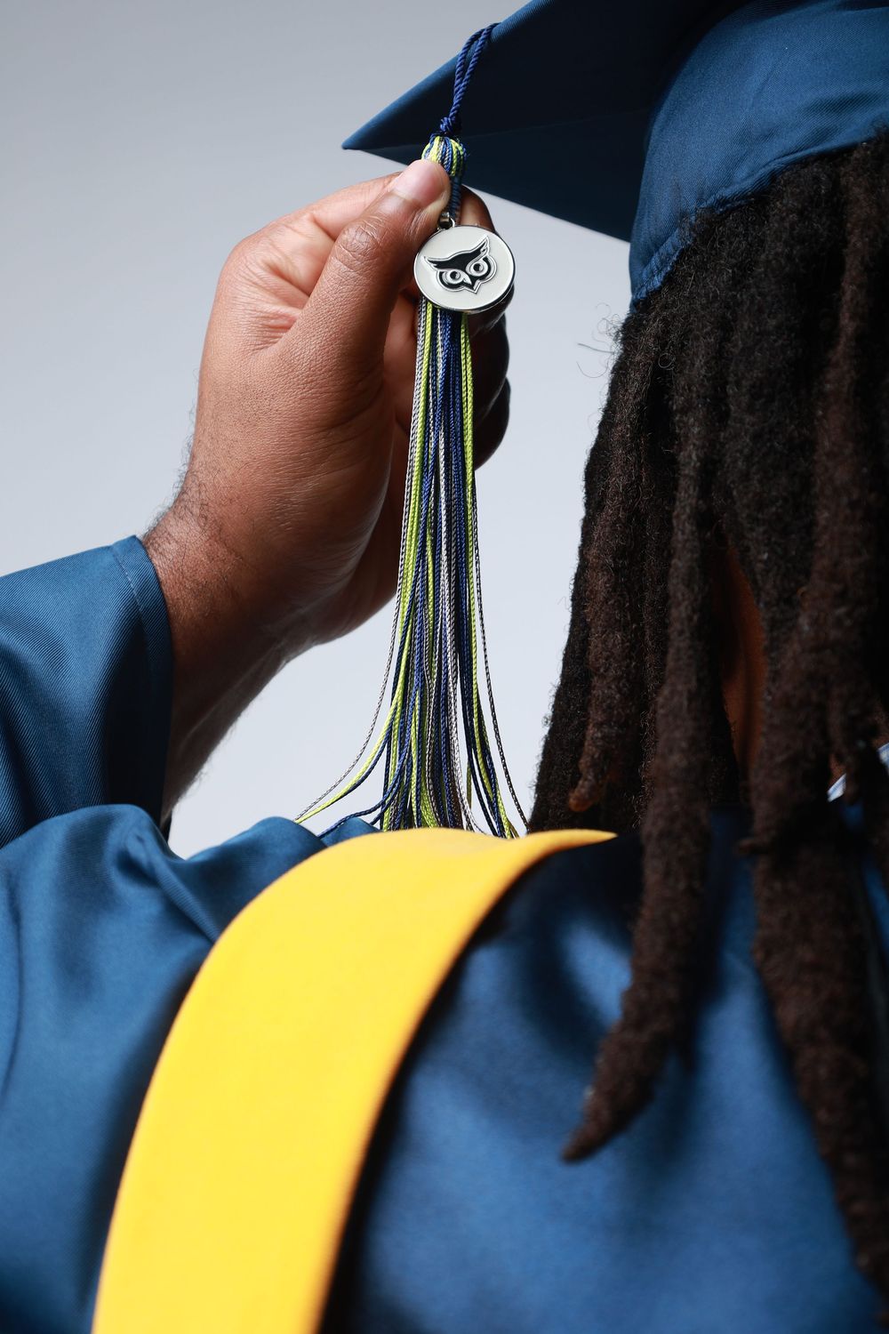 Graduate in blue cap and gown with dreadlocks holding tassel charm at graduation ceremony.