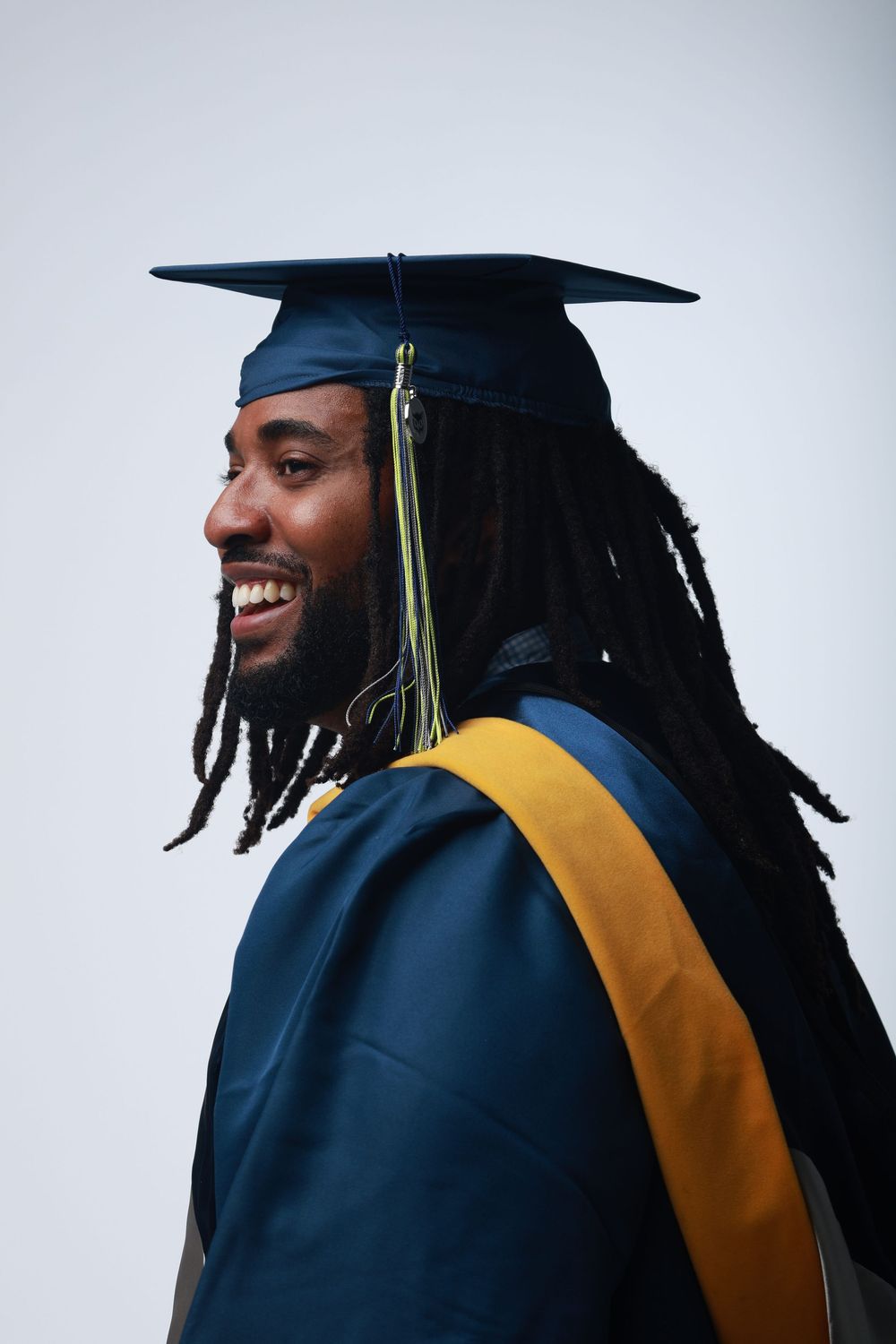 Smiling graduate in navy cap and gown with gold sash and dreadlocks, side profile view on light background.