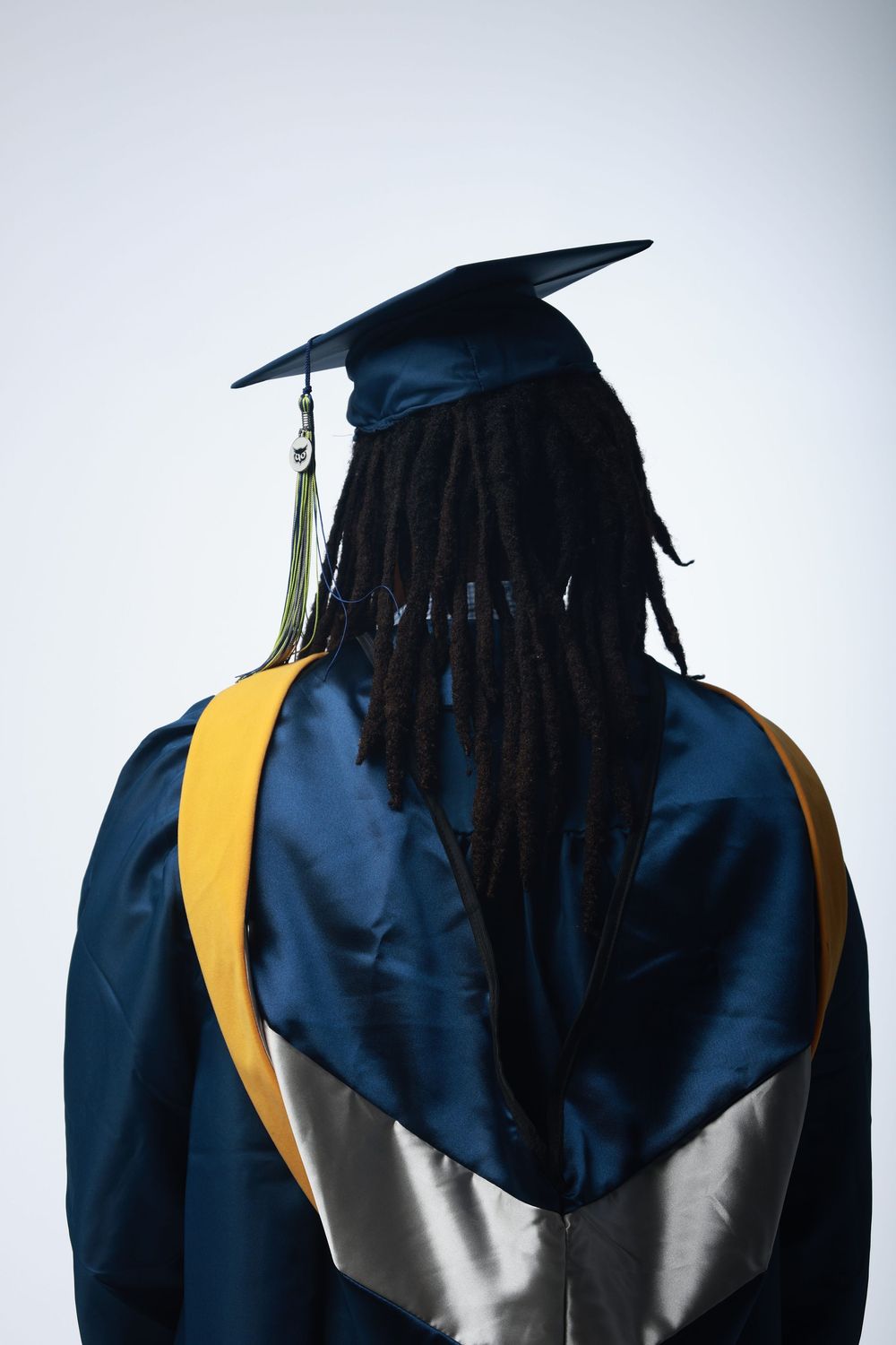 Graduate in navy blue cap and gown with gold hood, showing dreadlocks, viewed from behind.