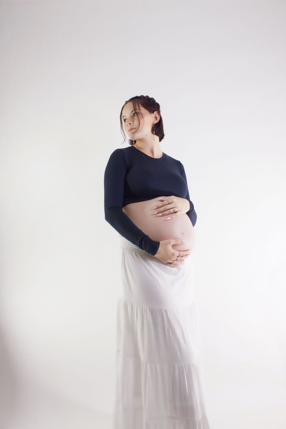 A pregnant woman in a navy blue top and white skirt poses sideways against a light backdrop while holding her belly.