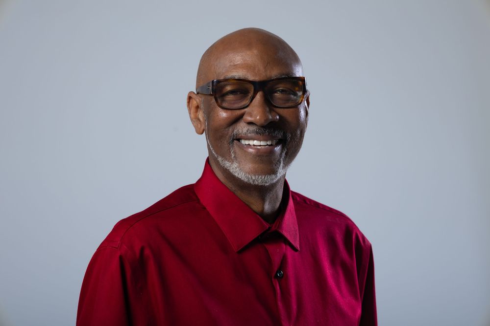 Smiling older man wearing glasses and a red shirt posed against a light gray background.