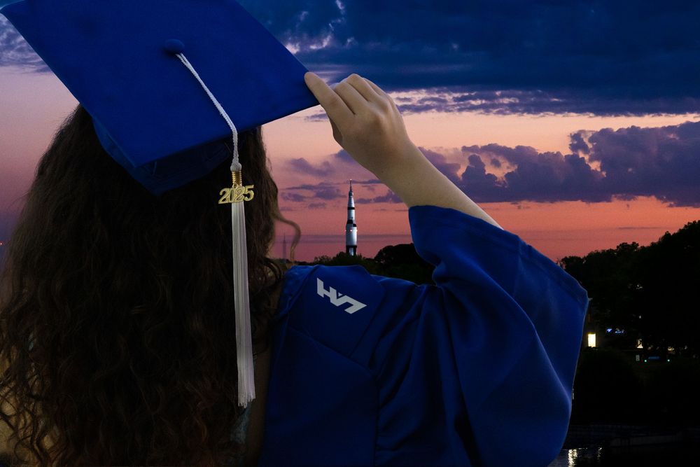Graduate in blue cap and gown holds tassel against vibrant sunset sky during outdoor graduation ceremony.