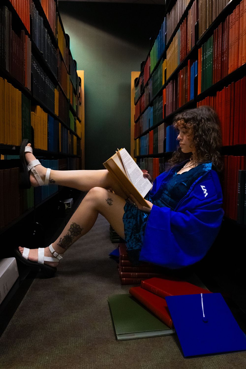 Someone reads casually in a bright blue jacket while sitting on the floor between library stacks with legs propped up.