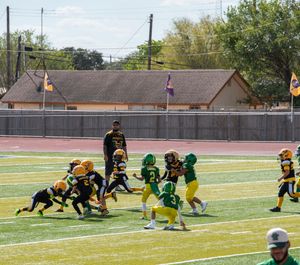 Youth football players in green and yellow uniforms compete during a game on an artificial turf field.