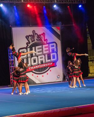Cheerleaders perform stunts at the Cheer World Championship competition on a blue mat under colorful stage lights.