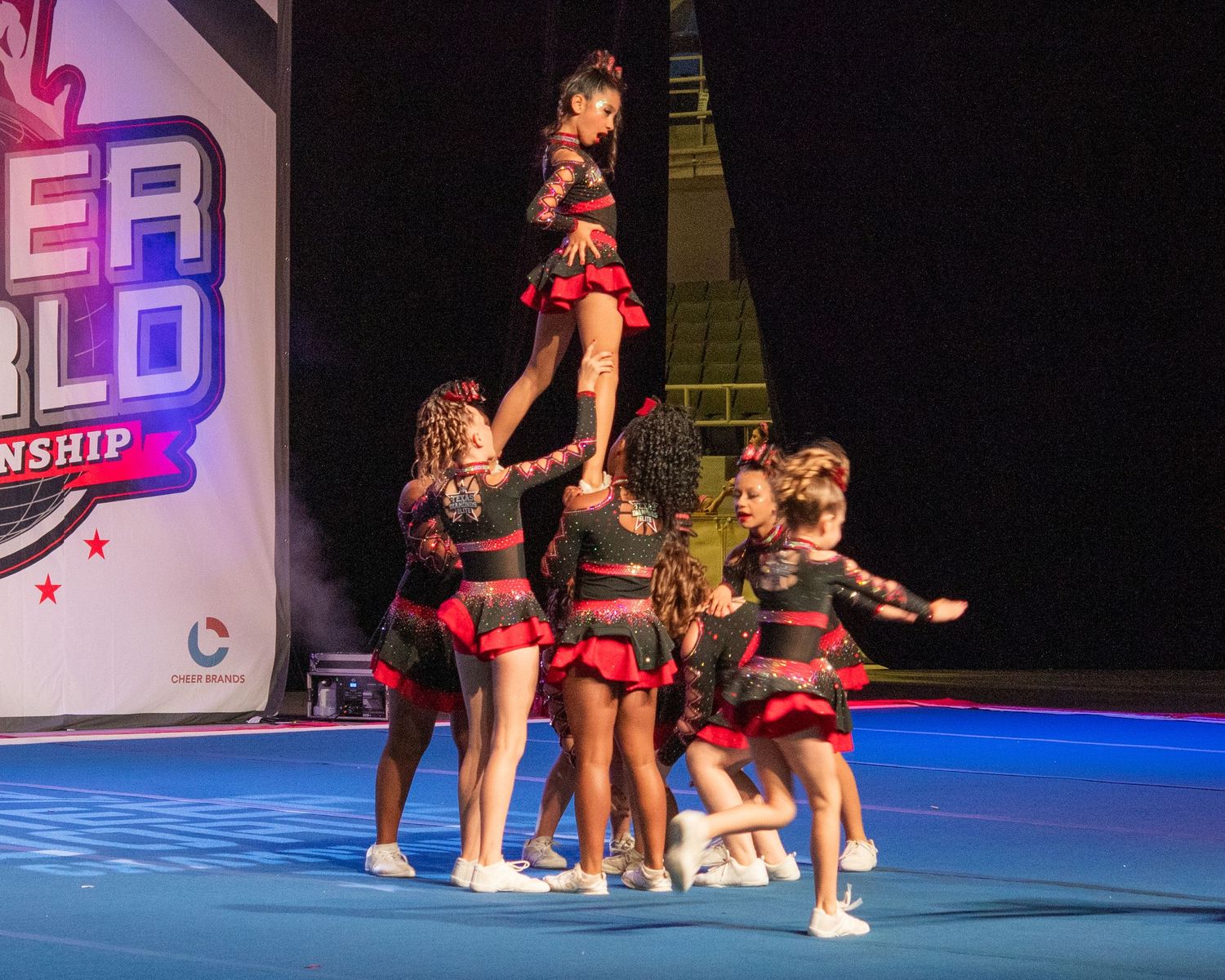 Cheerleaders perform a pyramid stunt in red and black uniforms at a Cheer World Championship competition.