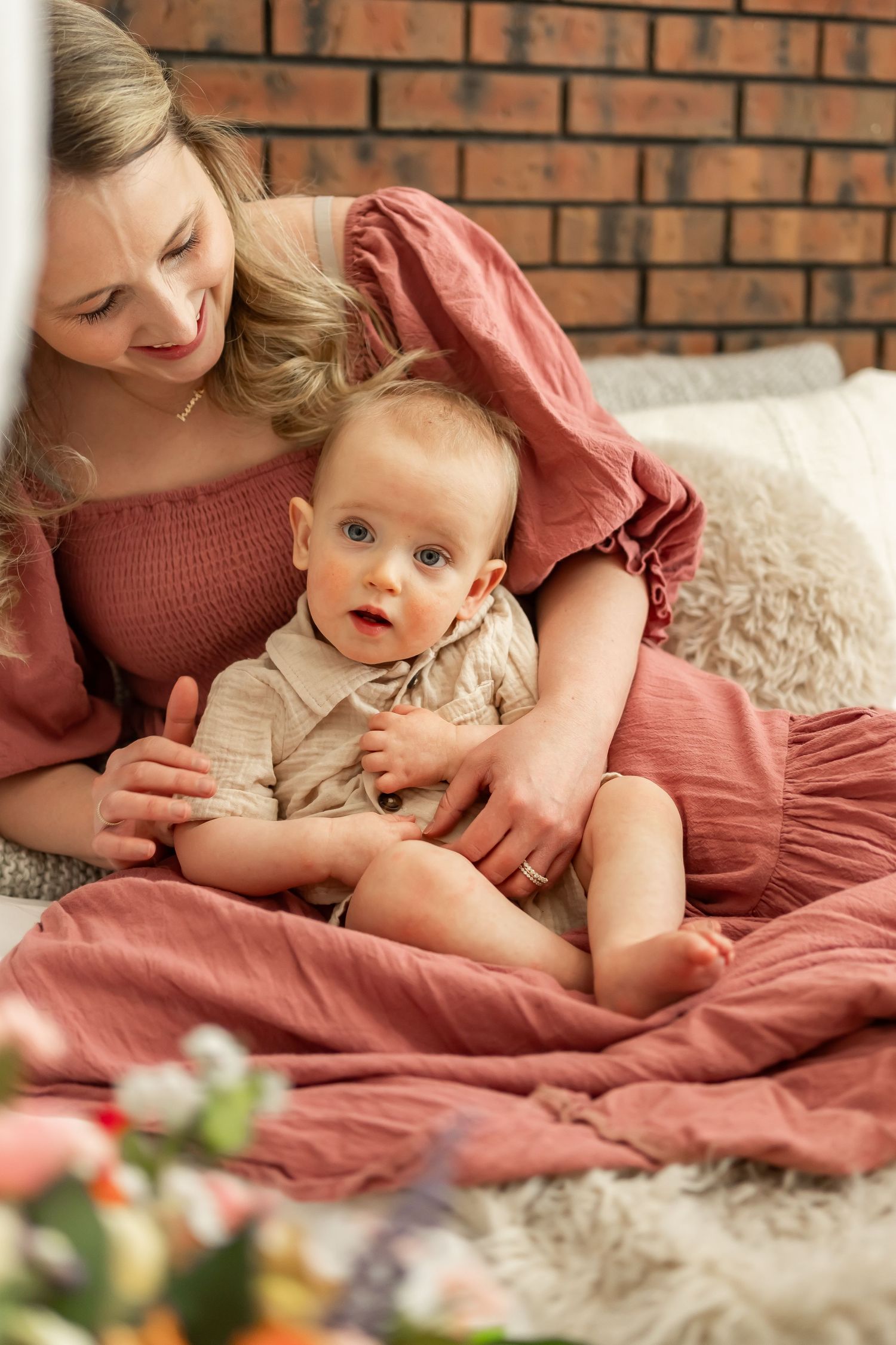 Mother in rose-colored dress cuddling with baby on cozy bed with brick wall background.