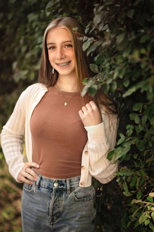 A teen wearing a brown ribbed tank top and cream cardigan poses outdoors against green foliage in natural lighting for senior photography