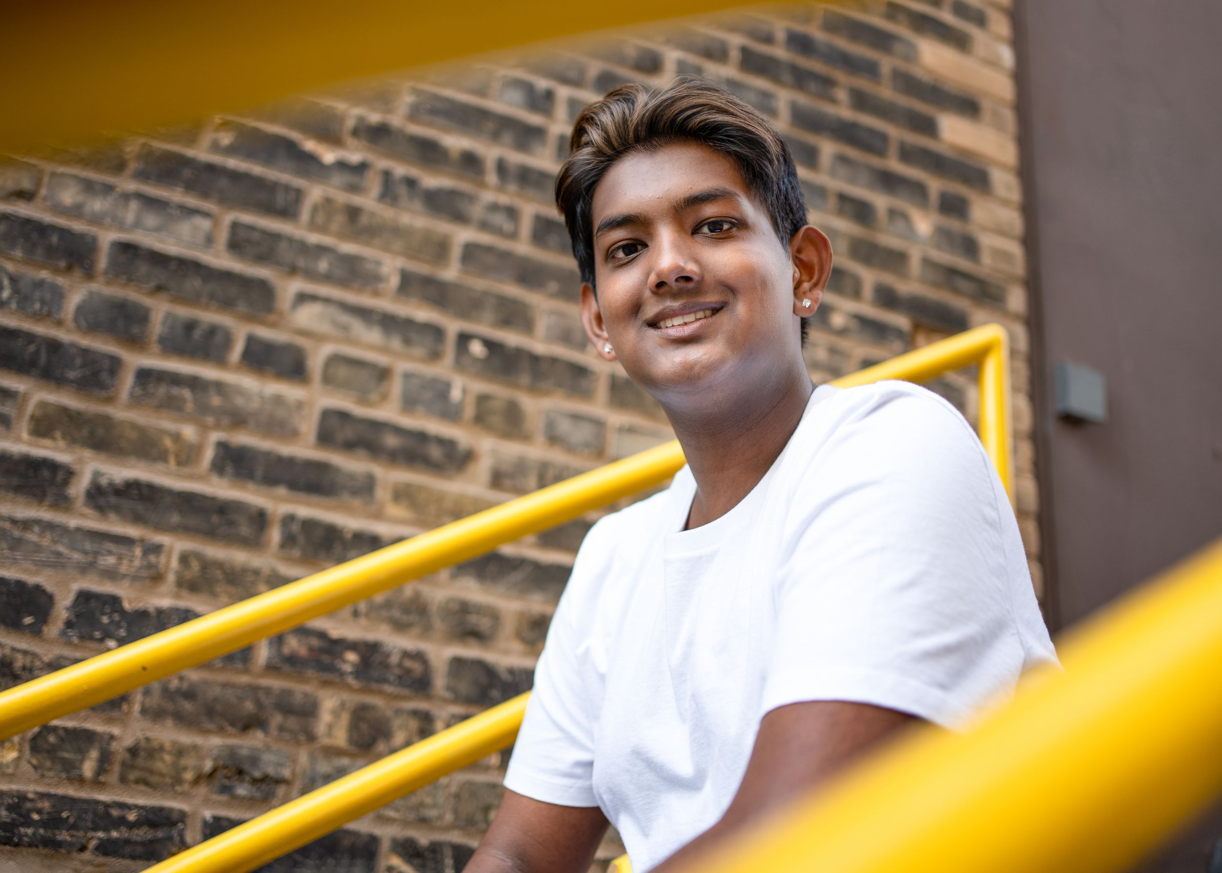 A person in a white t-shirt sits on yellow stairs against a brick wall, smiling warmly at the camera for senior photography in minneapolis