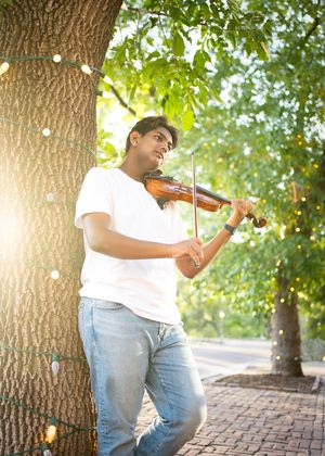 A musician plays violin while leaning against a tree trunk with string lights in a park setting for a minneapolis senior photographer