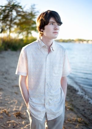 A person in a white button-down shirt poses on a beach at sunset with water and trees in the background for senior photos