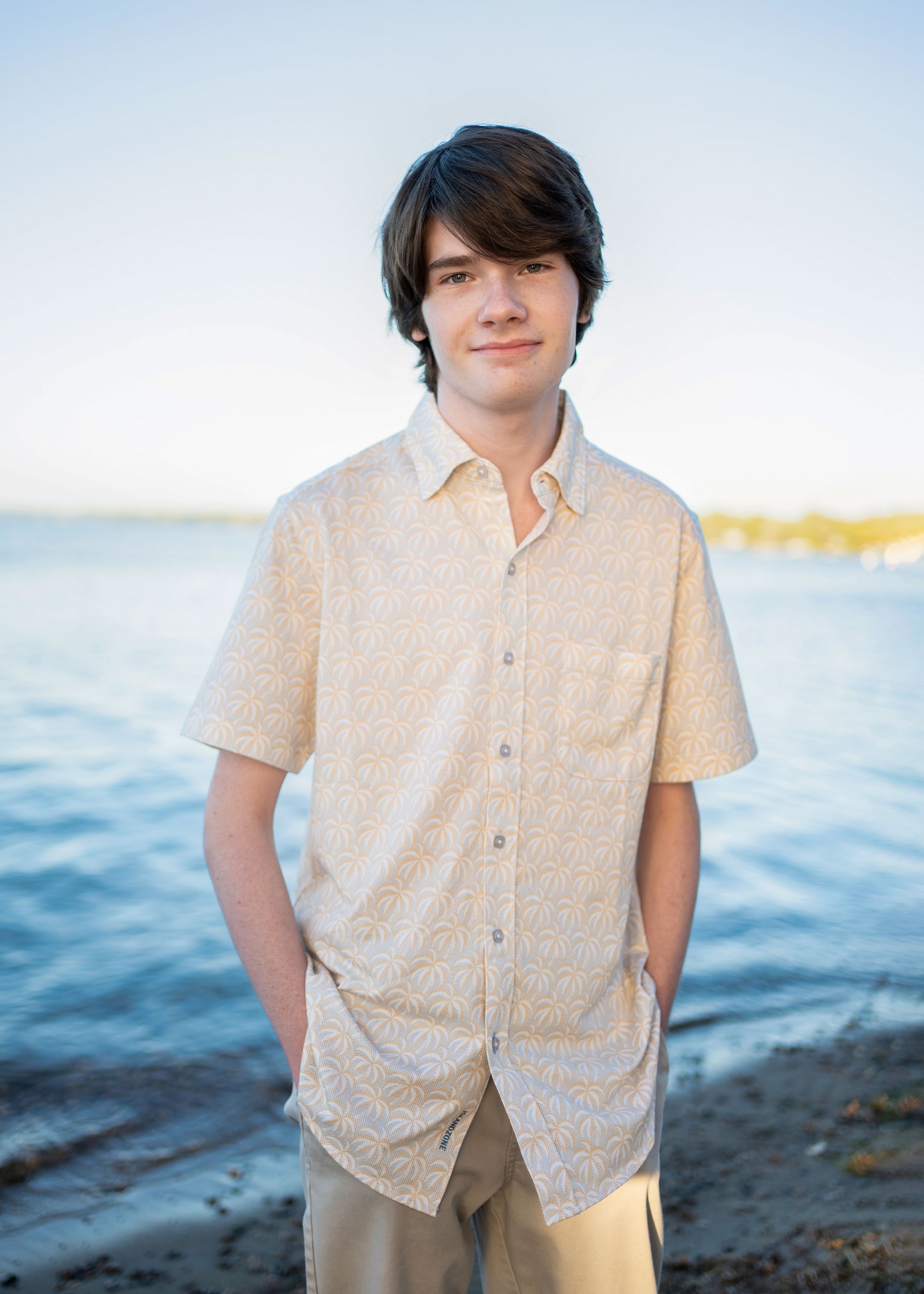 Teen in a beige button-down shirt stands by the waterfront on a sunny day with blue water in the background for minneapolis senior photos