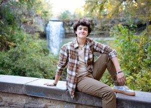 minneapolis senior photographer captures a teen in plaid shirt and khakis sitting on stone wall with scenic waterfall in background.