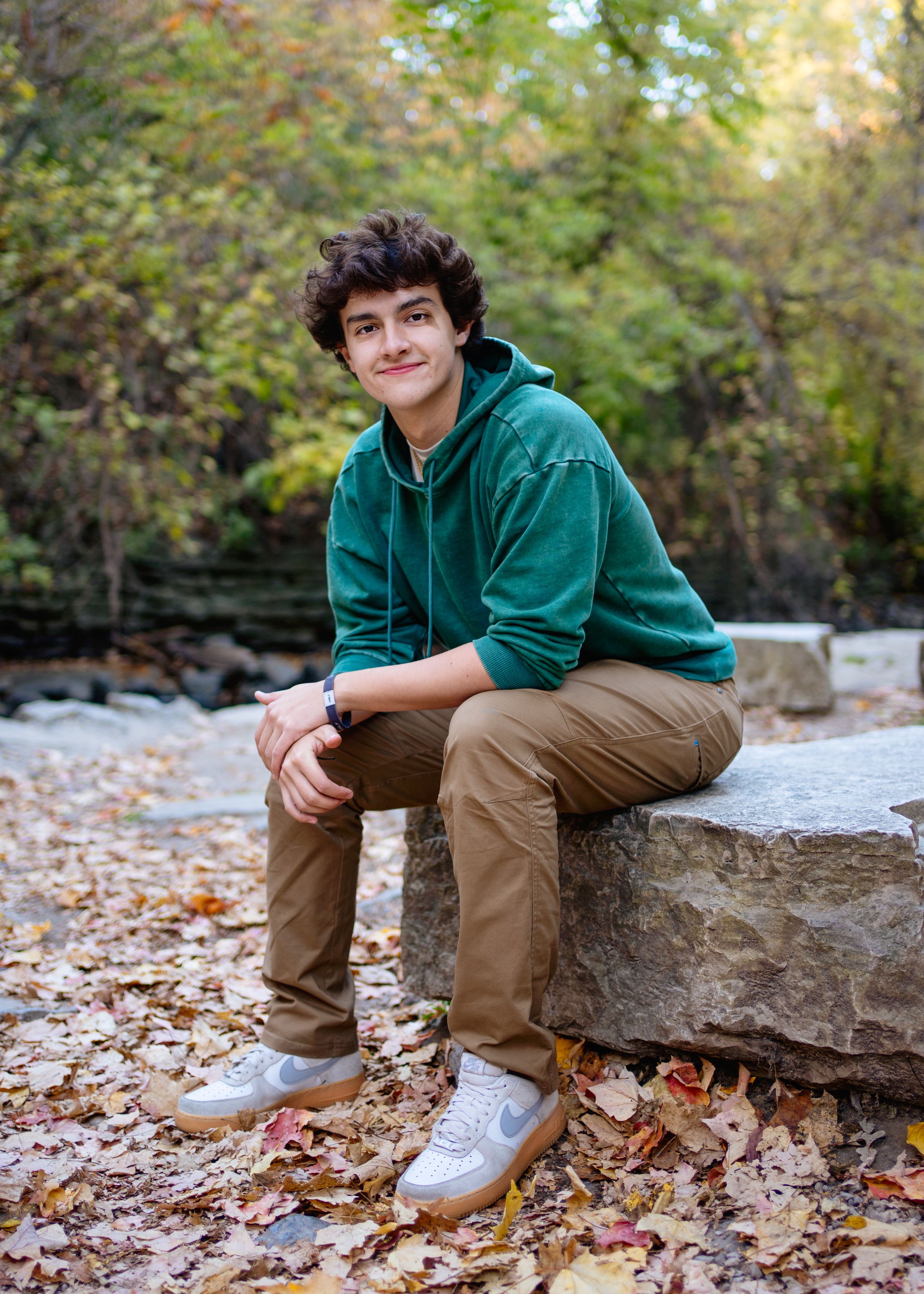 A minneapolis senior in a green fleece hoodie and khaki pants sits casually on a rock wall surrounded by autumn leaves in a wooded area.