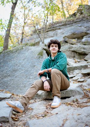 a teen relaxes on rocky outcrop wearing green hoodie and khaki pants during autumn hike on wooded trail for minneapolis senior photography