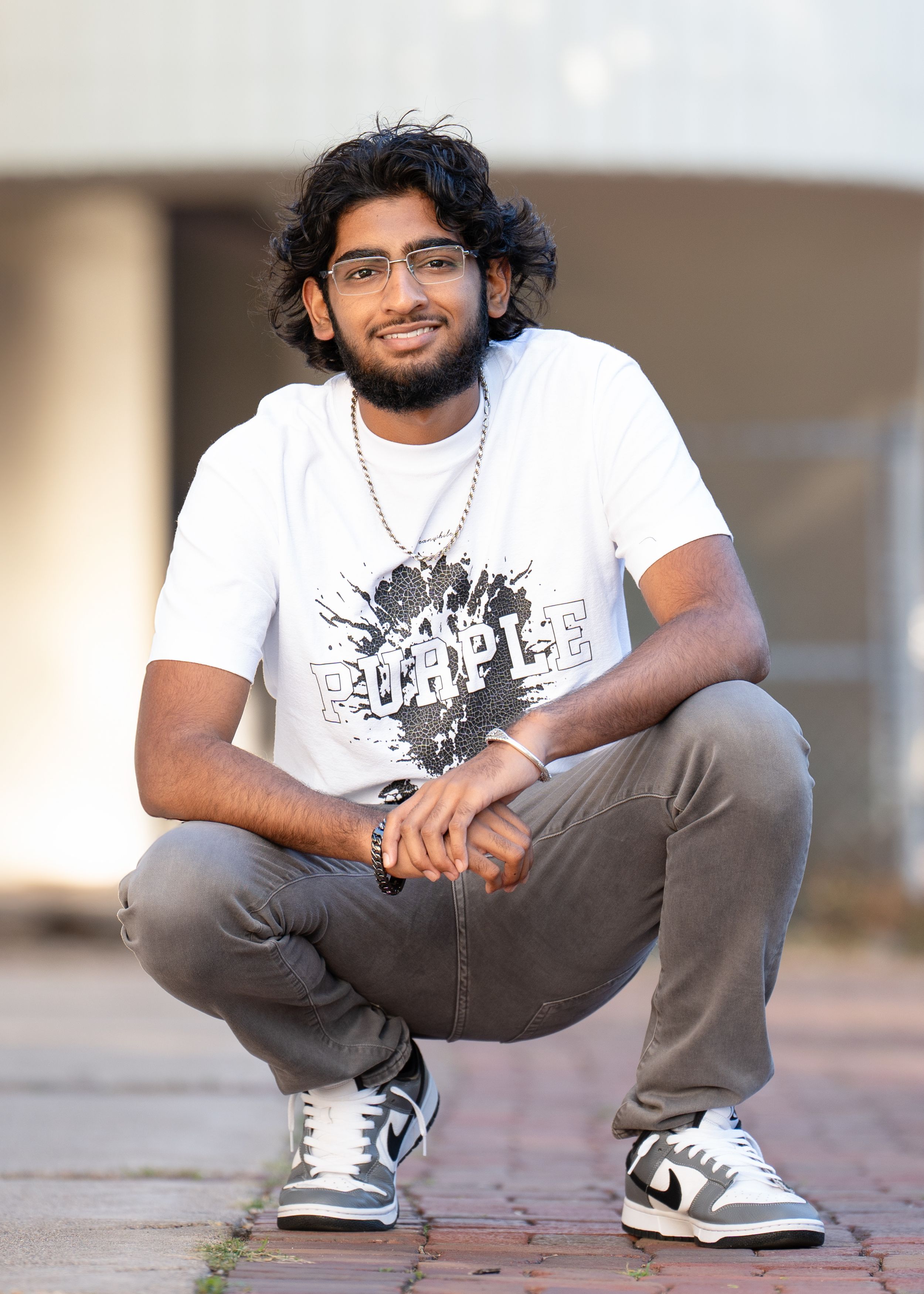 A person wearing glasses, a white graphic t-shirt and grey pants crouches casually on a brick pathway for senior photography in minneapolis