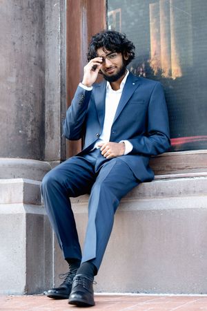 A teen in a navy blue suit sits on concrete steps wearing black dress shoes and touching their face for senior photographer in minneapolis