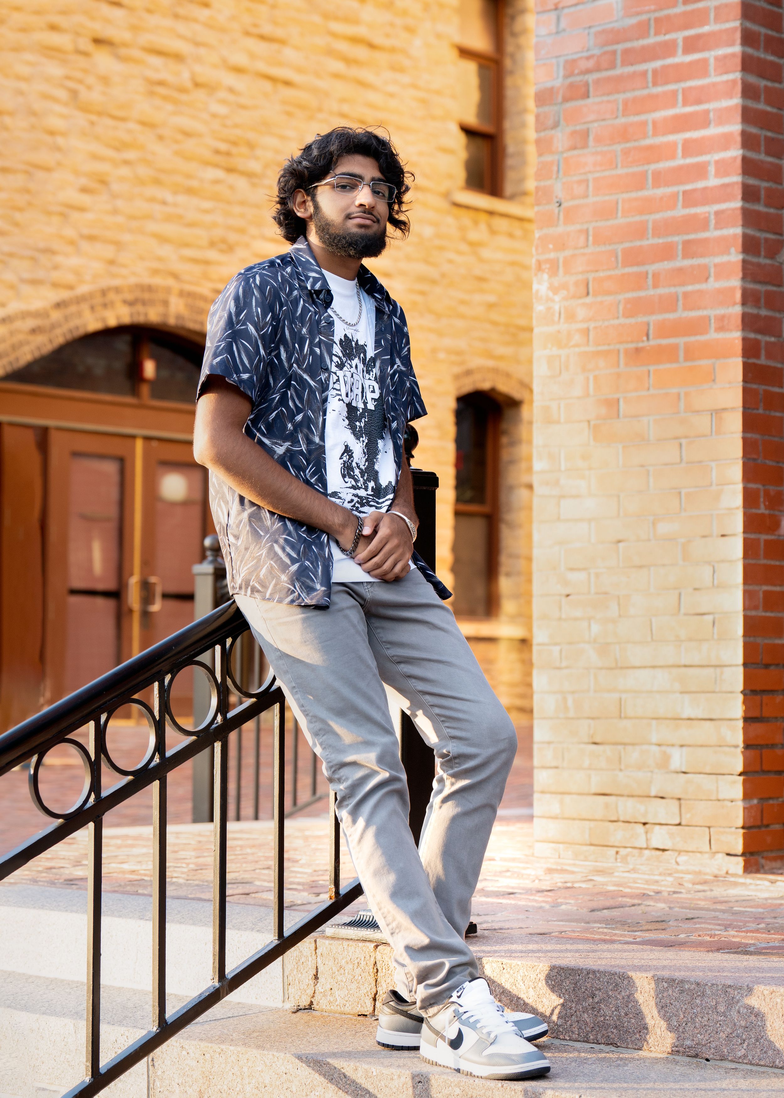 Casual model wearing patterned navy short sleeve shirt and gray pants poses against brick wall while leaning on railing for senior photos