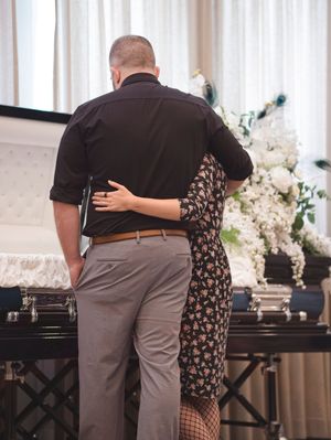 The son of the deceased and his wife stand in front of the casket, sharing a moment.