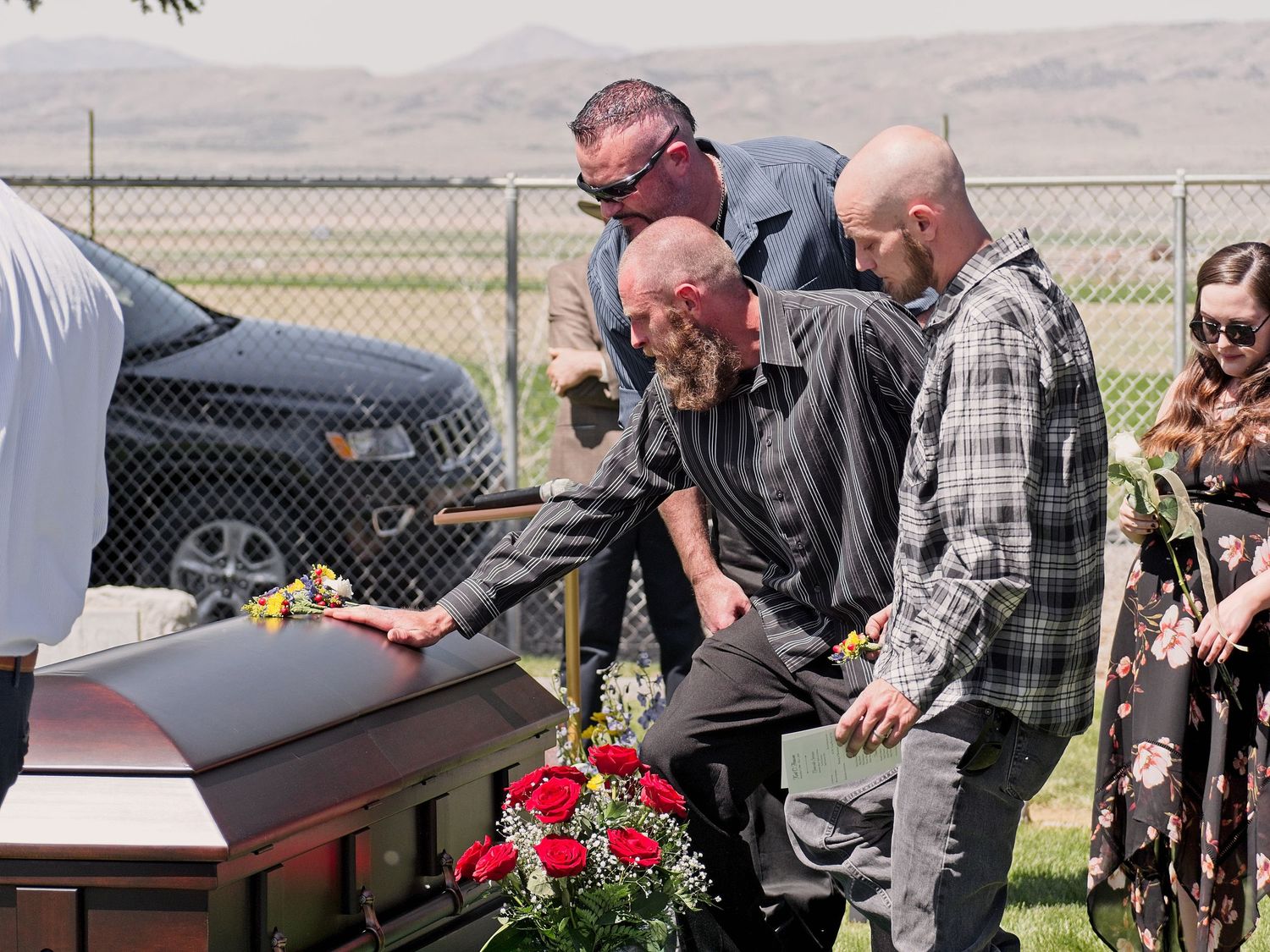 A man lays his hand on the top of the casket of his friend. Two others stand near him.