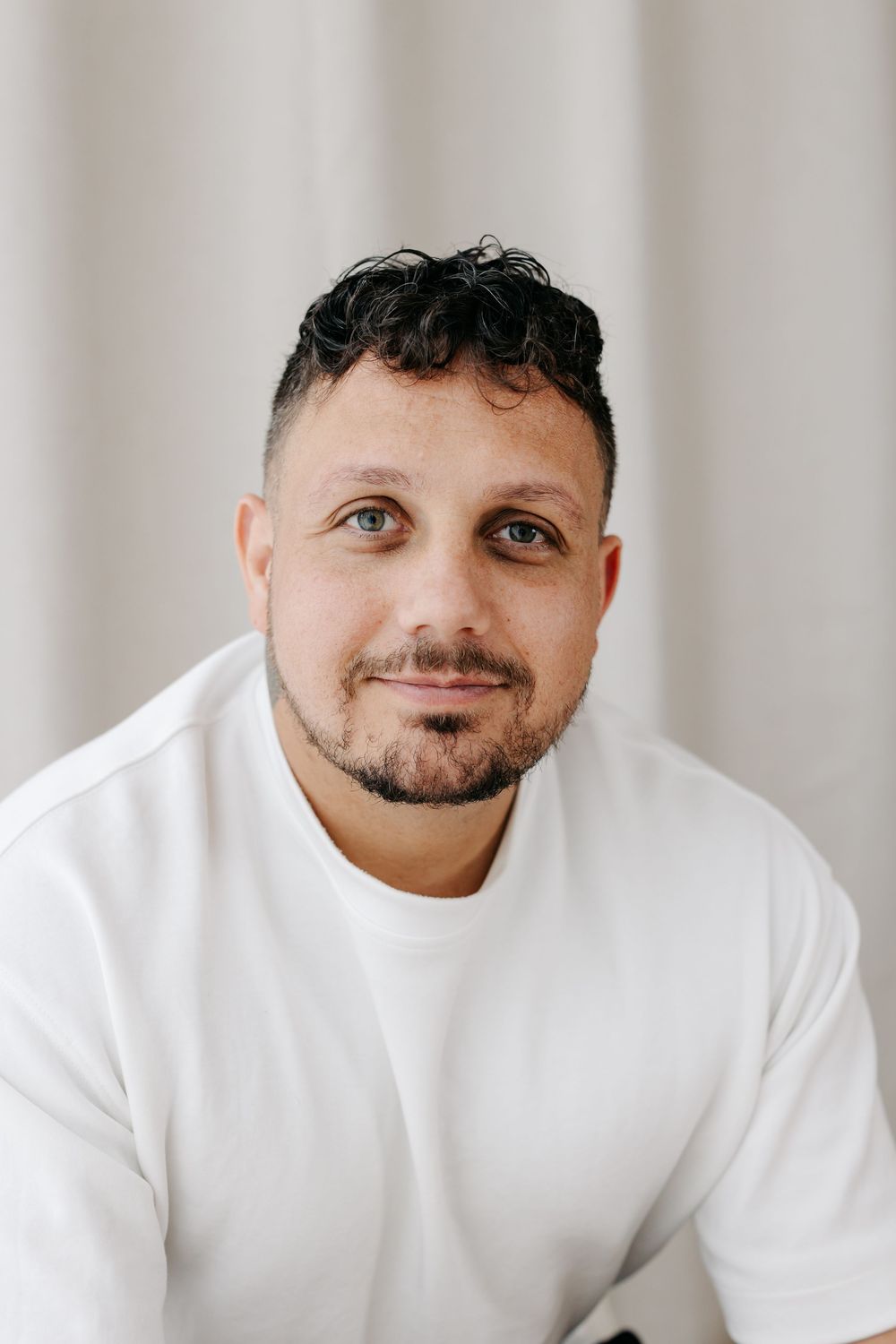 A man with dark curly hair and beard wearing a white t-shirt smiles softly against a light gray background.