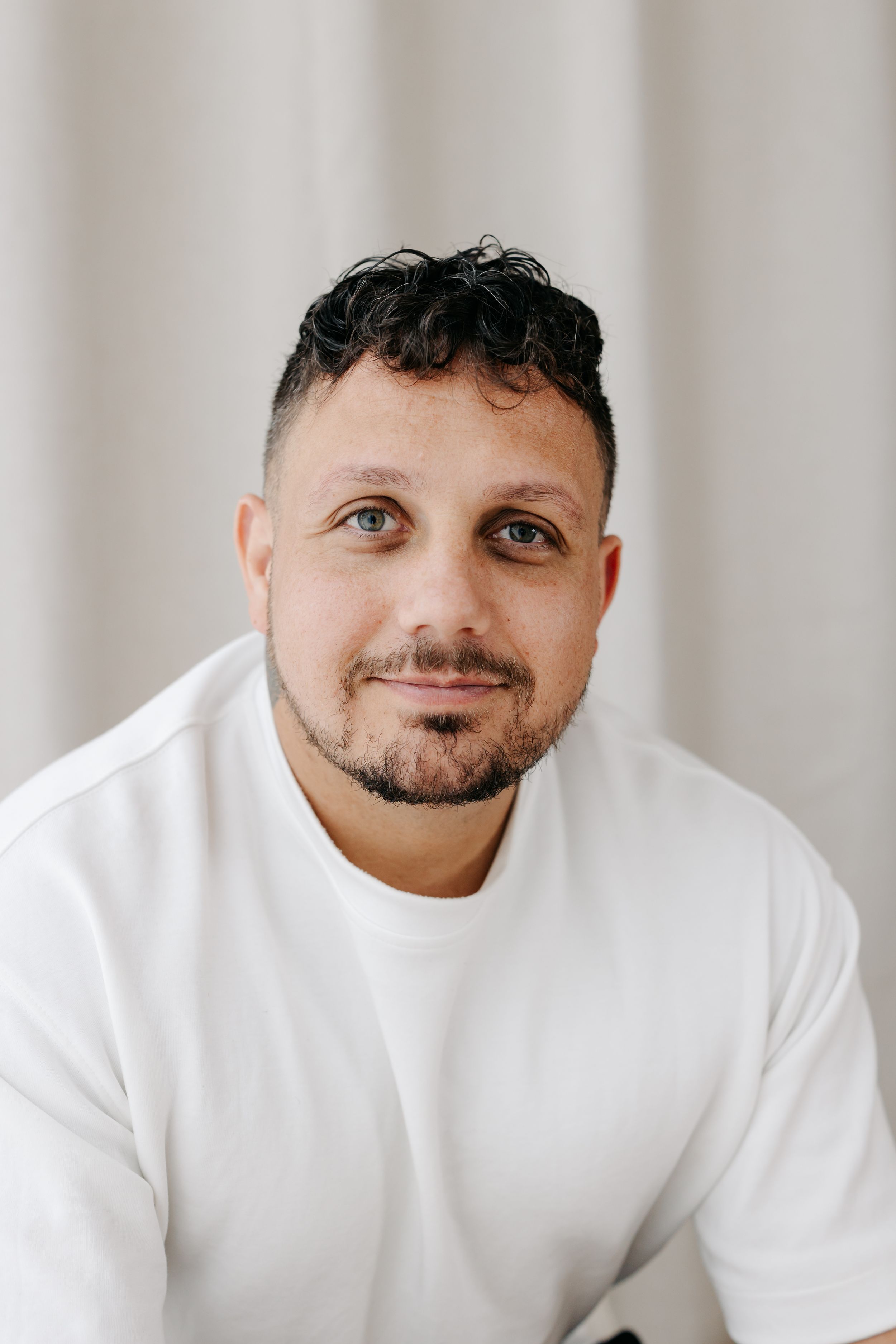 A man with dark curly hair and beard wearing a white t-shirt smiles softly against a light gray background.