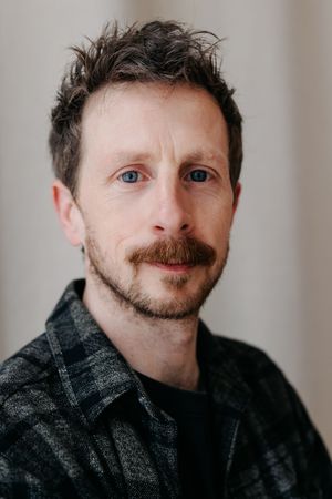 Man with blue eyes, mustache, and beard wearing a dark plaid shirt poses for a portrait photo.