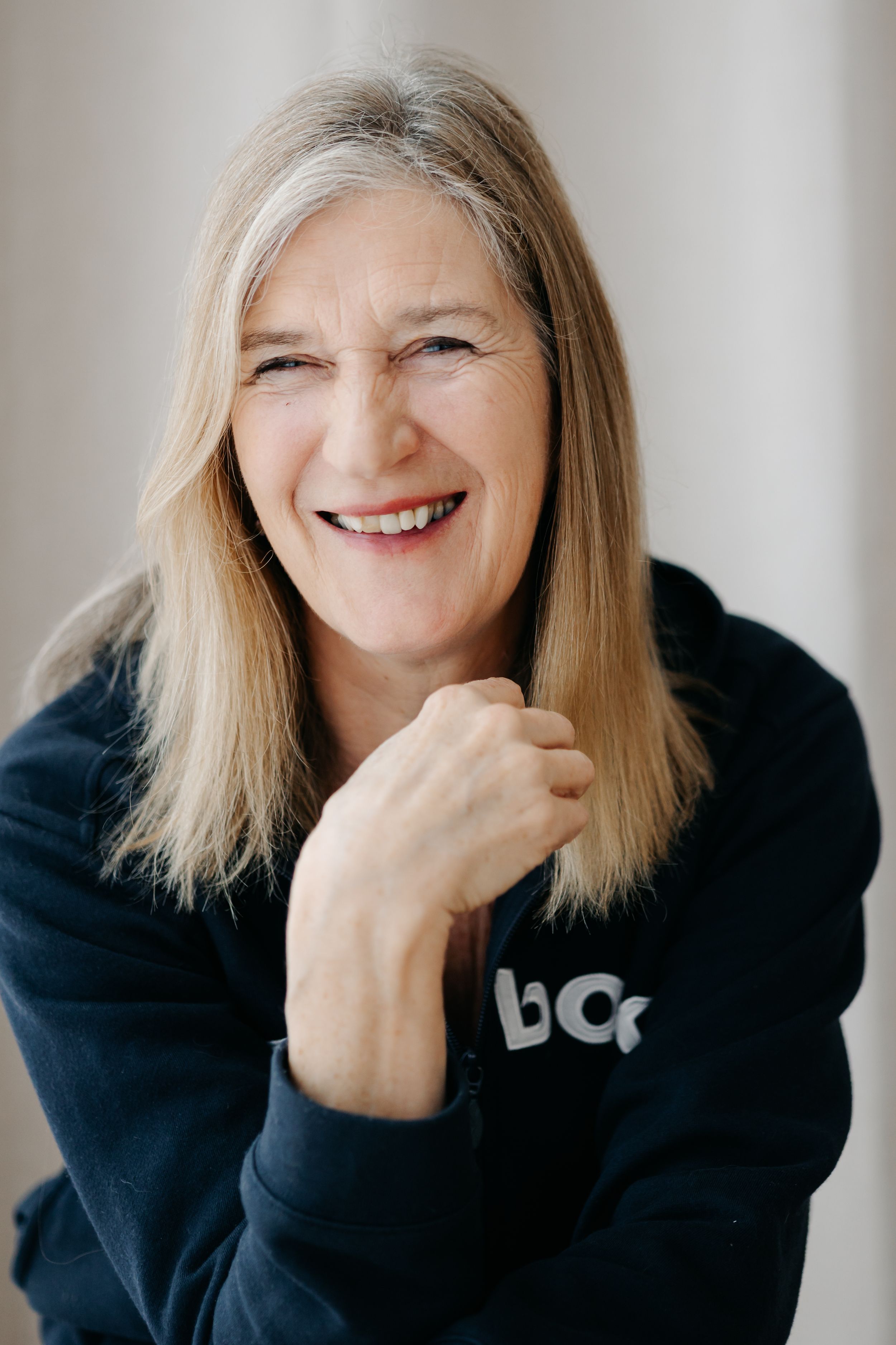 Smiling middle-aged woman with blonde hair wearing a dark navy hoodie, posing with hands clasped in a bright studio setting.