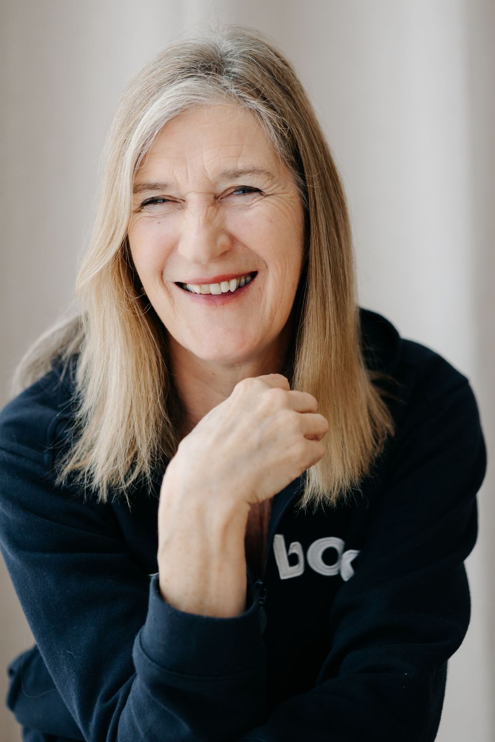 Smiling middle-aged woman with blonde hair wearing a dark navy hoodie, posing with hands clasped in a bright studio setting.