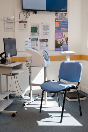 Medical waiting room with blue chair, health screening equipment, computer workstation, and informational posters on white wall.